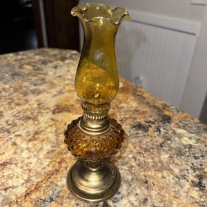Puede incluir: Lámpara de aceite de vidrio color ámbar con una parte superior con volantes y una base estampada. La lámpara tiene una base y detalles metálicos de color latón. La lámpara es de estilo vintage y probablemente se utiliza con fines decorativos.