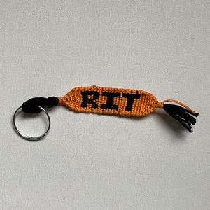 Può includere: Un portachiavi intrecciato arancione e nero con le lettere "RIT" in nero. Il portachiavi ha un nappa nera a un'estremità e un portachiavi argentato all'altra. Lo sfondo è una superficie bianca sporca.