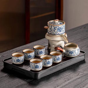 May include: A ceramic tea set with a blue floral pattern. The set includes a teapot, a serving pitcher, and six small teacups, all arranged on a dark wooden tray. The teapot sits on a white stand. The tea cups are filled with tea.