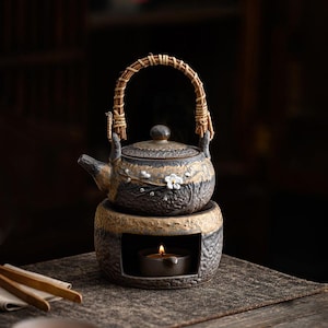 May include: A ceramic teapot with a woven handle, sitting atop a matching warmer with a candle inside. The teapot is decorated with white floral accents and a two-tone color scheme of brown and black. A tea set.
