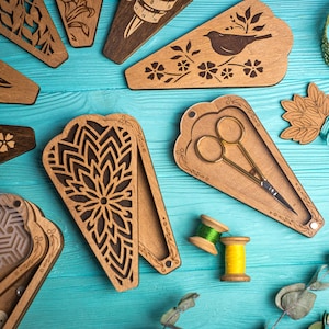May include: A collection of wooden needle cases with intricate laser-cut designs, including floral and geometric patterns. One case holds gold scissors. Two spools of thread are also visible, suggesting a sewing or crafting theme.