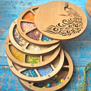 May include: Wooden craft organizer with multiple compartments filled with colorful beads. The organizer has a circular design with a lid featuring a peacock silhouette. The beads are in shades of orange, pink, white, yellow, green, and blue.