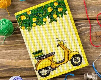 Op de afbeelding: Een gele kruissteek notitieboekhoes met een geel scooterontwerp. De bovenrand heeft een groen en geel citroenboomontwerp. De achtergrond is geel en wit gestreept. De hoes is omgeven door gele draad.