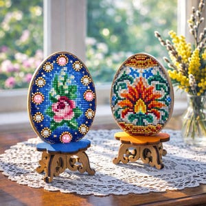 May include: Two decorative Easter eggs with detailed cross-stitch patterns. One shows a rose, the other a floral design. Each egg sits on a small, ornate wooden stand. The eggs are on a lace doily, with a vase of yellow flowers nearby.