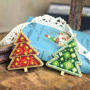 May include: Two wooden Christmas tree ornaments. One is red with yellow accents, the other is green with white and light green accents. Both have a beaded design and are on a wooden surface with a blue book and lace.