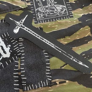 May include: A black fabric patch shaped like a war hammer with a white printed design of the weapon. The patch is positioned on a camouflage patterned fabric background alongside other black fabric patches with white stitched edges and graphics.