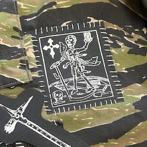 Puede incluir: Un parche rectangular de tela negra con gráficos serigrafiados en blanco de un esqueleto sosteniendo un reloj de arena, una cruz y una calavera, cosido sobre una tela de camuflaje con rayas de tigre en tonos verdes y marrones. Se ve otro parche parcial.