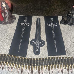 以下が含まれることがあります： 剣の白い線画が描かれた3枚の黒い布製パッチ。長方形のパッチ2枚が中央の剣の形をしたパッチを挟むように配置されています。手前には、真鍮と黒の金属製弾薬カートリッジが連なった長いベルトが置かれています。