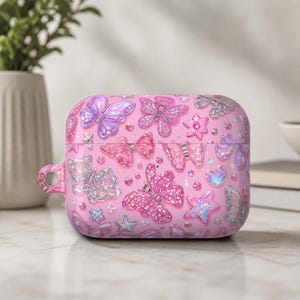 May include: A pink protective case for wireless earbuds featuring a repeating pattern of iridescent butterflies, sparkling stars, and small heart shapes. The surface has a glossy finish with various shades of pink, purple, and silver accents.