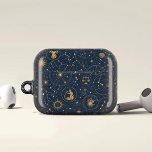 May include: A dark blue protective case for wireless earbuds featuring a gold celestial pattern with various zodiac signs, constellations, and stars. The case has a small circular loop on the left side and is flanked by two white wireless earbuds.
