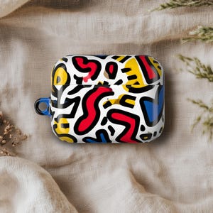 May include: A white AirPod case with a vibrant abstract pattern in red, yellow, blue, and black. The case has a blue loop and is displayed on a neutral fabric surface.