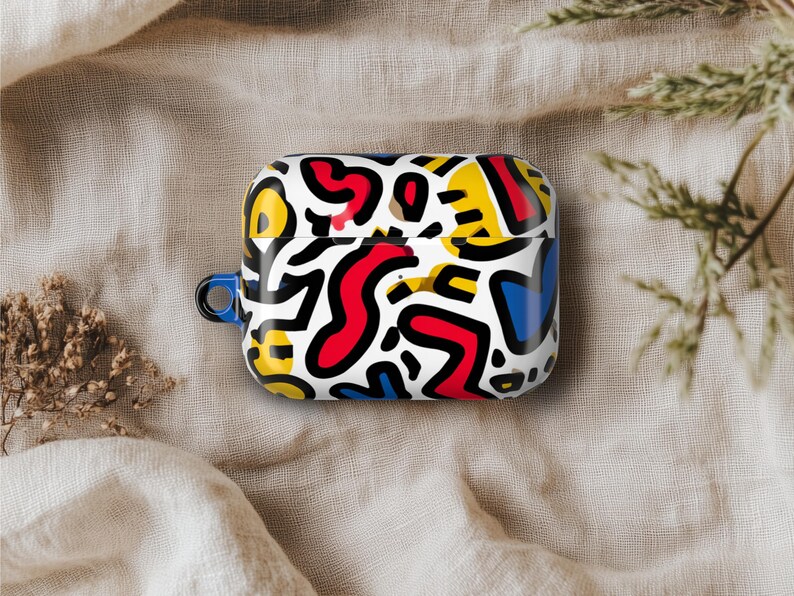 May include: A white AirPod case with a vibrant abstract pattern in red, yellow, blue, and black. The case has a blue loop and is displayed on a neutral fabric surface.