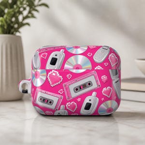 May include: A bright pink protective case for wireless earbuds featuring a repeating pattern of retro technology including silver flip phones, compact discs, clear cassette tapes, and small pixelated hearts scattered across the surface.