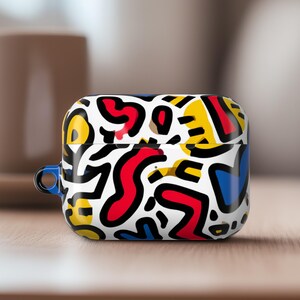 May include: A white AirPod case with a colourful abstract pattern in red, yellow, blue, and black. The case has a blue loop and sits on a wooden surface.
