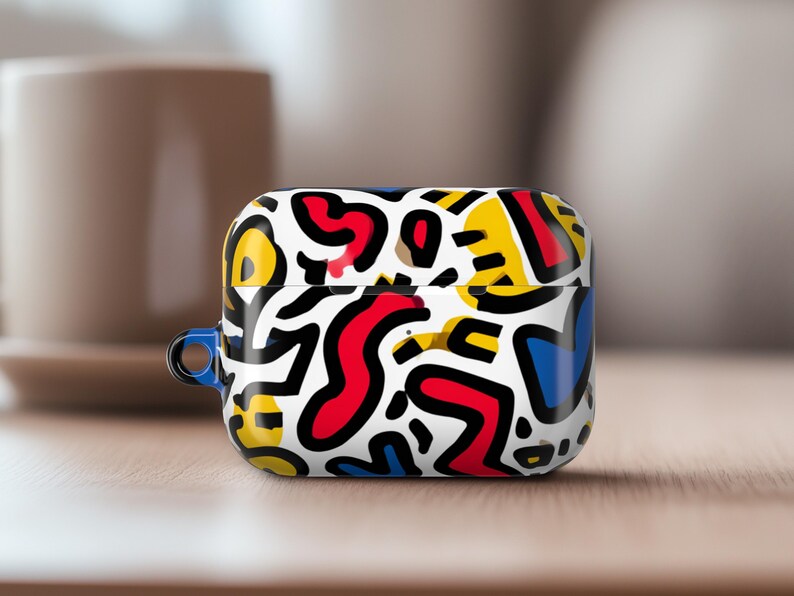 May include: A white AirPod case with a colourful abstract pattern in red, yellow, blue, and black. The case has a blue loop and sits on a wooden surface.