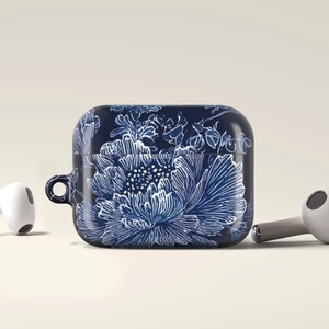 May include: A navy blue AirPods case with a white floral design, featuring a large peony. Two white and gray wireless earbuds are positioned on either side of the case. The case has a small loop for attaching to a keychain.