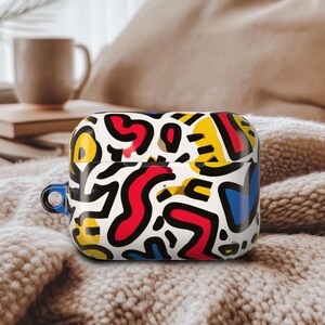 May include: A white AirPod case with a vibrant abstract pattern in red, yellow, blue, and black. The case has a blue loop. It rests on a soft, textured surface. The design is eye-catching and modern.