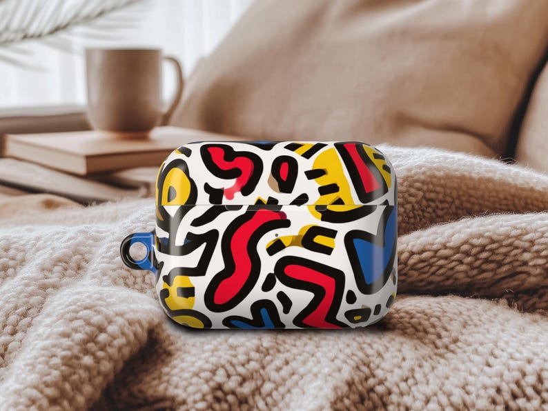 May include: A white AirPod case with a vibrant abstract pattern in red, yellow, blue, and black. The case has a blue loop. It rests on a soft, textured surface. The design is eye-catching and modern.