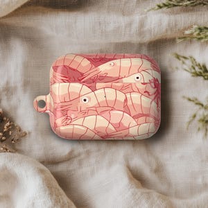 May include: A rectangular, pink and beige AirPod case featuring a shrimp illustration. The case has a small loop on the side and is set against a neutral background with dried plants.