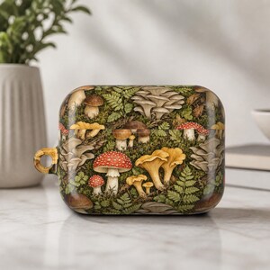 May include: A rectangular protective case for wireless earbuds featuring a detailed botanical print of various mushrooms, including red-capped toadstools and yellow chanterelles, nestled among green ferns and mossy textures with a side attachment loop.