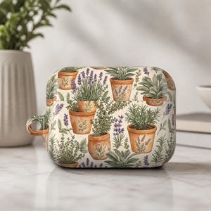 May include: A rectangular protective case for wireless earbuds featuring a repeating pattern of various green herbs and purple lavender sprigs planted in small terracotta-colored pots against a light cream-colored background.
