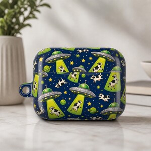 May include: A dark blue protective case for wireless earbuds featuring a repeating pattern of silver UFOs with bright green tractor beams lifting black and white cows, interspersed with yellow stars and small green planets across the surface.