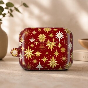 May include: A rectangular protective case for wireless earbuds featuring a deep red background with a repeating pattern of gold and white multi-pointed stars. The case includes a small circular loop on the left side for attaching a lanyard or clip.