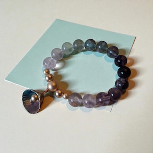 May include: A beaded bracelet featuring a gradient of purple and gray stones. The bracelet includes a seashell charm with a pearl accent and smaller gold-colored beads. The bracelet is displayed on a light blue surface.