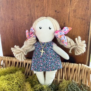 May include: A fabric doll with light beige yarn hair styled in two braids tied with pink and blue floral bows. The doll wears a dark blue dress with a small pink and blue floral pattern and a gold-toned necklace featuring a cross pendant.