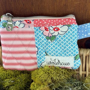 May include: A rectangular quilted zipper pouch featuring a patchwork design of pink horizontal stripes and a blue floral pattern. A white fabric label with the black text watercue is stitched to the front. The pouch has a silver metal zipper.