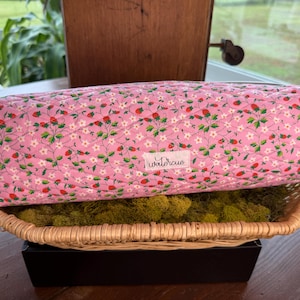 May include: A pink quilted pencil case with a strawberry and floral pattern. The case is rectangular and has a zipper closure. It rests on a woven basket filled with green moss.