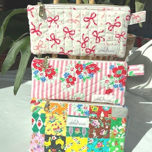 May include: Three quilted fabric pouches with zipper closures. The top pouch is white with red bows, the middle one has red flowers and pink stripes, and the bottom one is a patchwork of colorful patterns. Each pouch has the word "watercue" on it.
