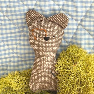 May include: A small, stuffed fabric animal toy made from brown and white checkered cloth. It features two pointed ears, embroidered black eyes, a black nose, and a decorative orange stitched detail above one eye. The toy has a simple, flat shape.