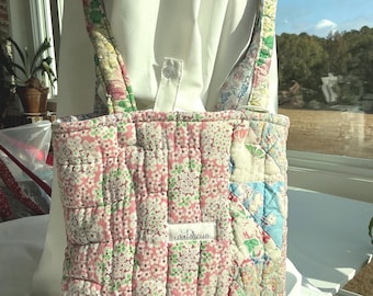 May include: A quilted tote bag with a floral pattern in pink, white, and green. The bag features a patchwork design with a variety of floral fabrics. The straps and trim have a floral pattern. The label reads "watercue".