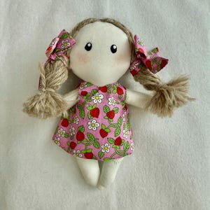 May include: A fabric doll with light skin, brown yarn hair styled in two pigtails, and pink blush on the cheeks. The doll wears a pink dress featuring a repeating pattern of red strawberries, green leaves, and white flowers, with matching bows.