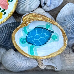 May include: Hand-painted seashell trinket dishes. One dish features a blue turtle design, another has a floral pattern. Both have gold painted rims. The dishes rest on a bed of seashells.