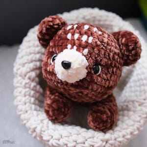 May include: A handmade brown plush bear with white spots on its head and a white muzzle. The bear has black eyes and is nestled in a cream-colored crocheted basket. The plush toy is soft and cuddly.