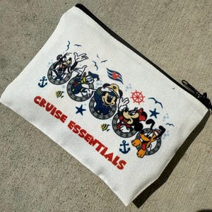 May include: White canvas zipped pouch with a black zip. The pouch features a nautical design with cartoon characters in portholes, anchors, stars, and the text "CRUISE ESSENTIALS" in red.