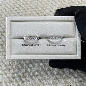 May include: Two diamond eternity rings displayed in a white box. The text above the rings reads "5 Stones Eternity" and "11 Stones Eternity". The rings are set against a textured white background.