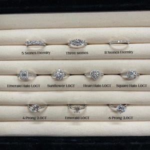 May include: A display of various diamond rings, including eternity bands and rings with emerald, heart, and square halo settings. The rings are set in a black tray with text labels such as "5 Stones Eternity" and "4 Prong 2.0CT".