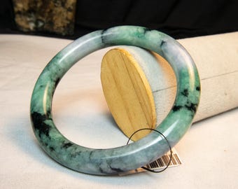 May include: A jade bangle bracelet with a smooth, polished surface. The bracelet features a mix of green and black hues, with a translucent quality. The bangle is displayed on a wooden stand.
