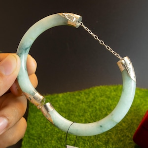 May include: A light green jade bangle bracelet with silver-colored metal accents. The bracelet is held open by a delicate chain. The jade has a marbled appearance with darker green veining. The bracelet is displayed against a green and black background.