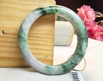 Op de afbeelding: Een jade armband met een gemarmerd uiterlijk, met tinten groen en wit. De gladde, cirkelvormige armband wordt gepresenteerd tegen een houten standaard en een wit oppervlak, met een aangehecht label.