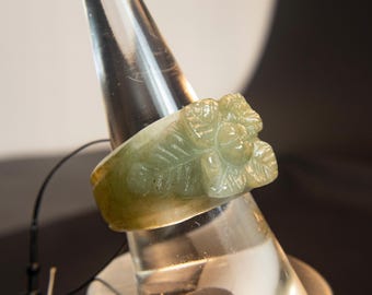 以下が含まれることがあります： 彫刻された花のディテールが施された、明るい緑色の翡翠の指輪。指輪は透明な円錐形のリングホルダーに展示されています。指輪のデザインは葉と花を特徴とし、翡翠の自然な色のバリエーションを際立たせています。