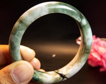 Op de afbeelding: Een jade armband met een gemarmerd uiterlijk. De armband heeft een mix van witte en groene tinten, met donkerdere groene adering. Het gladde, gepolijste oppervlak reflecteert licht en benadrukt de natuurlijke variaties in de steen.