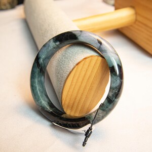 Puede incluir: Un brazalete de jade con un patrón veteado en verde oscuro y negro. El brazalete se exhibe sobre un soporte cilíndrico gris claro con un soporte de madera. El brazalete tiene forma circular y parece pulido.