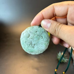 May include: A round, pale green jade pendant with a carved dragon design. The pendant is suspended from a dark green cord with yellow accents. The jade has a translucent quality, and the dragon is intricately detailed.