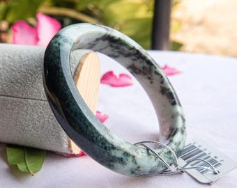 May include: A jade bangle bracelet with a marbled appearance, featuring shades of green and white. The smooth, polished surface reflects light, showcasing the natural stone's unique patterns. The bracelet is displayed on a light-colored surface, with a tag attached.