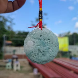 May include: A round, pale green jade pendant with a detailed dragon carving. The pendant is suspended by a red and black cord. The background is a blurred view of a blue sky and greenery.