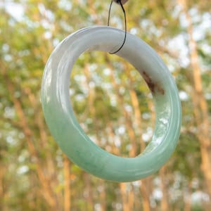 Puede incluir: Un brazalete de jade con un degradado de color, que va de un tono pálido, casi blanco, a un verde suave. La superficie lisa y pulida refleja la luz, mostrando las variaciones naturales de la piedra. Se observa una pequeña mancha oscura.